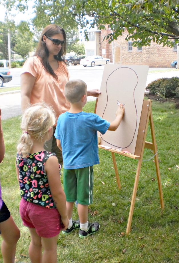 [photo of two children playing a game at an easle, with an adult standing by]