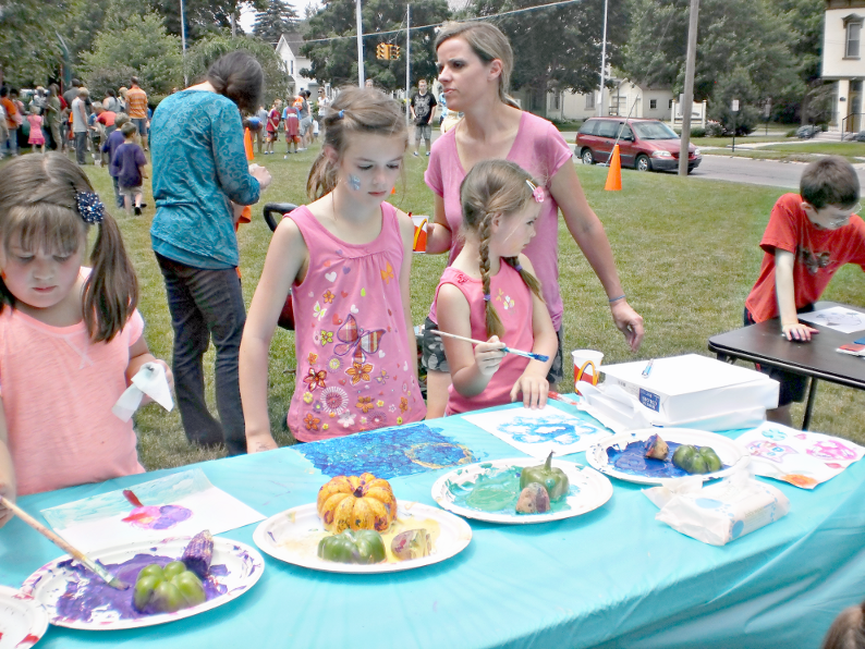 [photo of three girls painting at a table, with a crowd of people milling about on the lawn behind them]