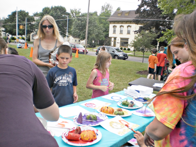 [photo of two children looking at the artwork on the painting table, with several adults standing around]